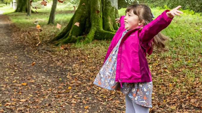 A girl in a pink coat looks happy as she throws fallen leaves into the air